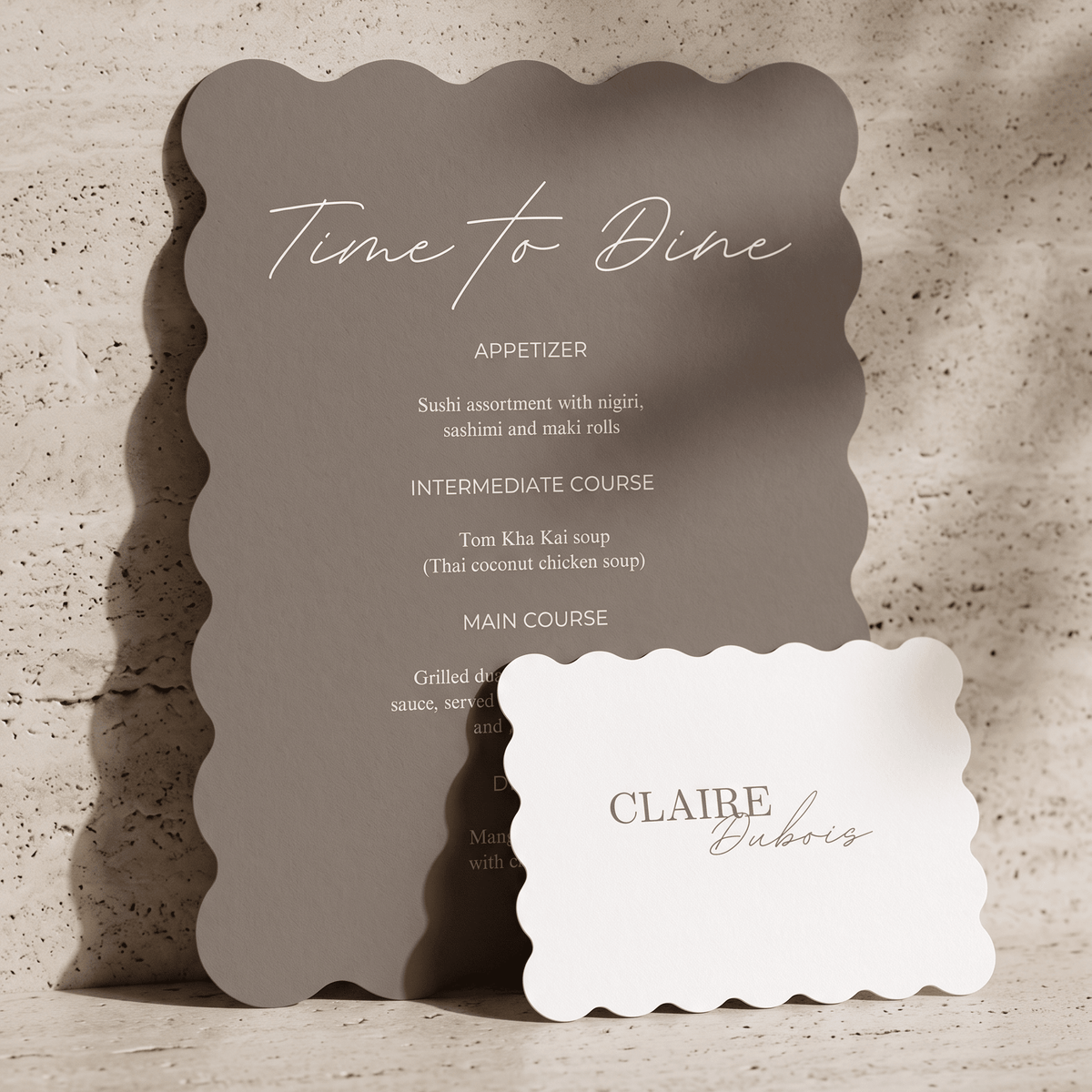 A scalloped-edge dinner menu card in anthracite with white script text reading 'Time to Dine' and a separate white name card reading 'CLAIRE Babes,' styled on a textured beige surface with soft shadows.