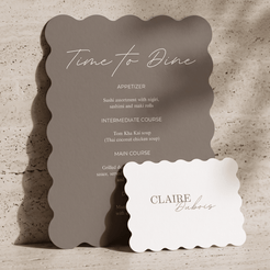 A scalloped-edge dinner menu card in anthracite with white script text reading 'Time to Dine' and a separate white name card reading 'CLAIRE Babes,' styled on a textured beige surface with soft shadows.