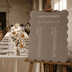 A scalloped gray wedding seat chart sign on an easel, displayed in a beautifully decorated wedding reception hall with a long table set in the blurred background.