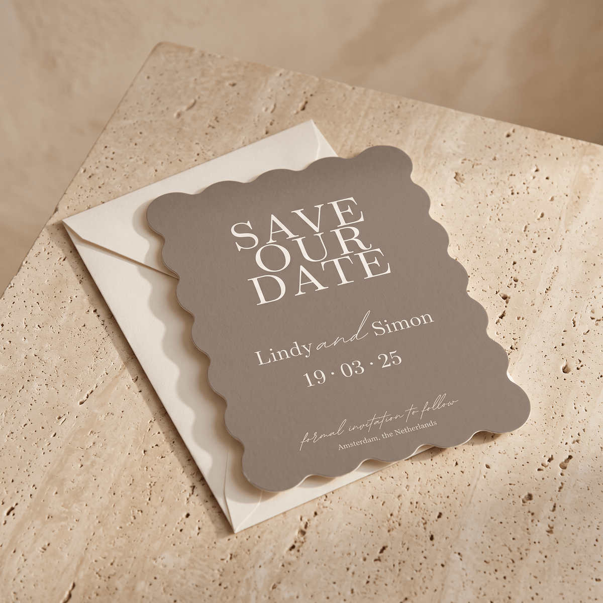A scalloped-edge 'Save Our Date' card in antracite with white lettering, placed on top of a cream envelope over a beige stone surface.