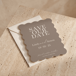 A scalloped-edge 'Save Our Date' card in antracite with white lettering, placed on top of a cream envelope over a beige stone surface.
