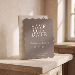 A scalloped-edge gray 'Save Our Date' card with white text stands in front of a white envelope on a stone ledge in a sunlit interior.