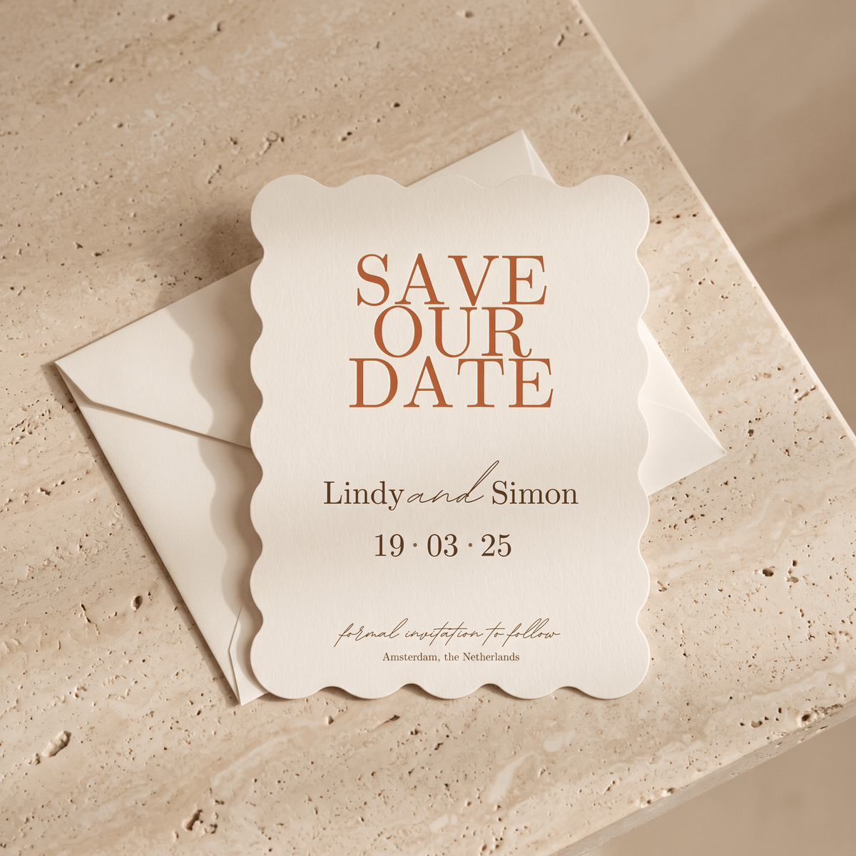 A scalloped cream wedding save-the-date card with terracotta lettering reading 'SAVE OUR DATE' and names/date details, placed on top of a matching envelope on a beige stone surface.