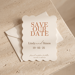 A scalloped cream wedding save-the-date card with terracotta lettering reading 'SAVE OUR DATE' and names/date details, placed on top of a matching envelope on a beige stone surface.