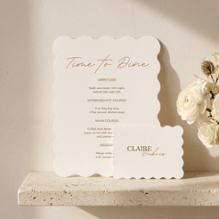 A scalloped cream paper menu card with elegant copper-gold typography reading 'Time to Dine' and course details, styled on a stone ledge beside white flowers against a warm beige wall.
