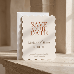 A scalloped cream wedding save-the-date card with terracotta lettering reading 'SAVE OUR DATE' and names/date details, layered over a white envelope on a stone surface in warm natural light.