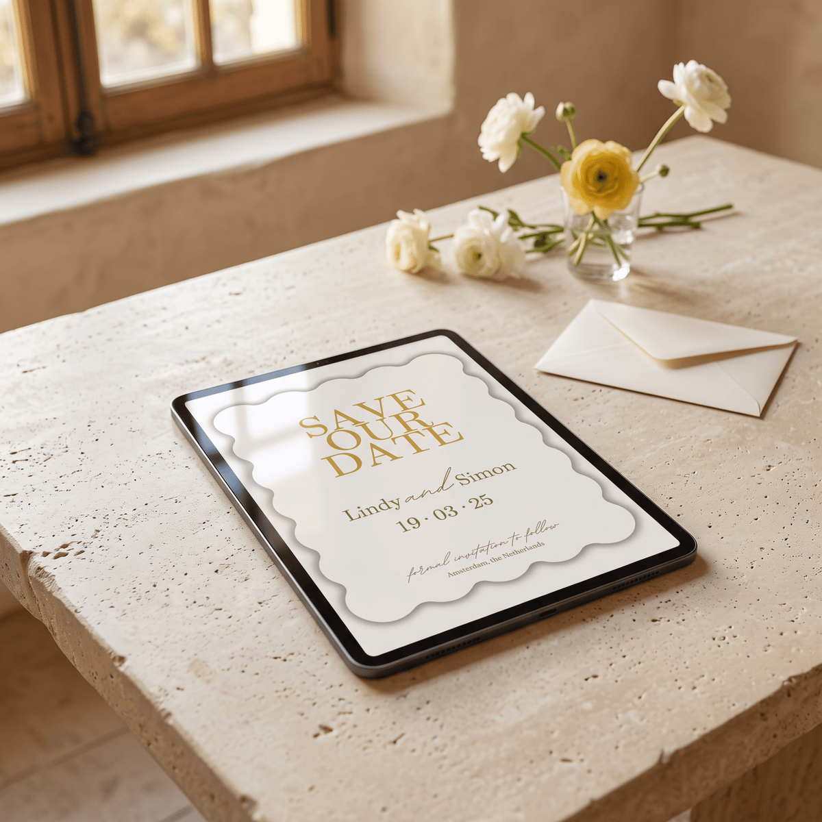 A tablet on a stone table displaying a wedding save-the-date card design, with cream envelopes and small white and yellow flowers nearby in a softly lit interior setting.