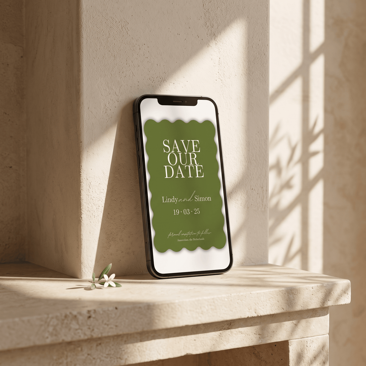 A smartphone leaning against a beige stone wall on a matching stone ledge, displaying an olive green 'Save Our Date' card with white text. The scene is lit by warm natural sunlight with soft shadows and a small white flower on the ledge.