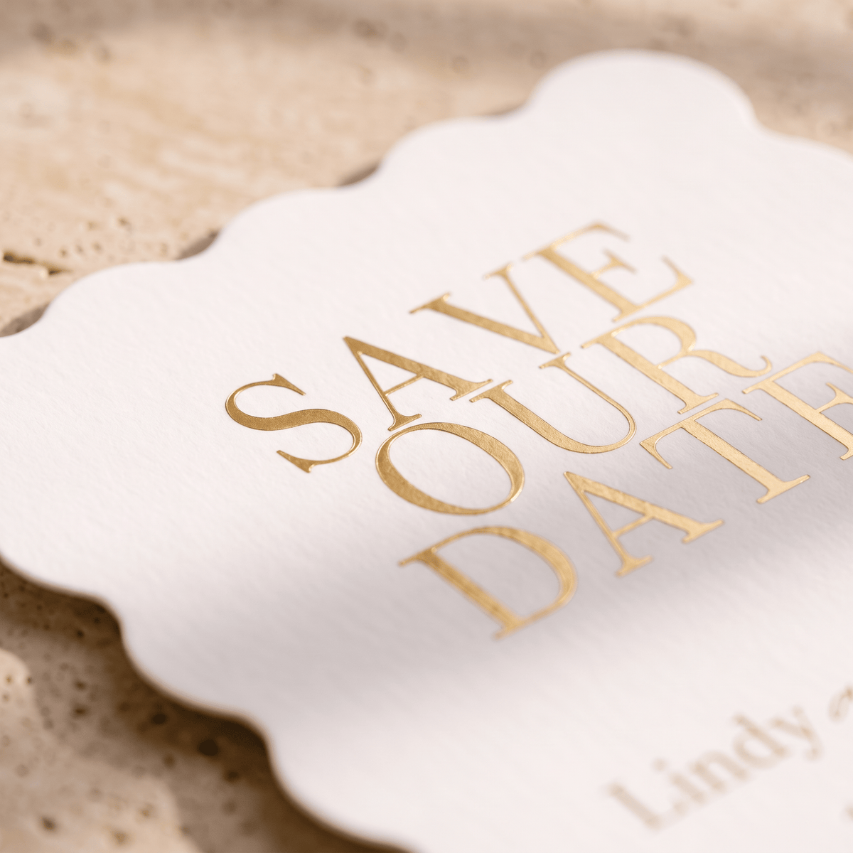 A close-up of a white scalloped paper card with elegant gold foil lettering that reads 'SAVE OUR DATE' on a textured beige stone surface.