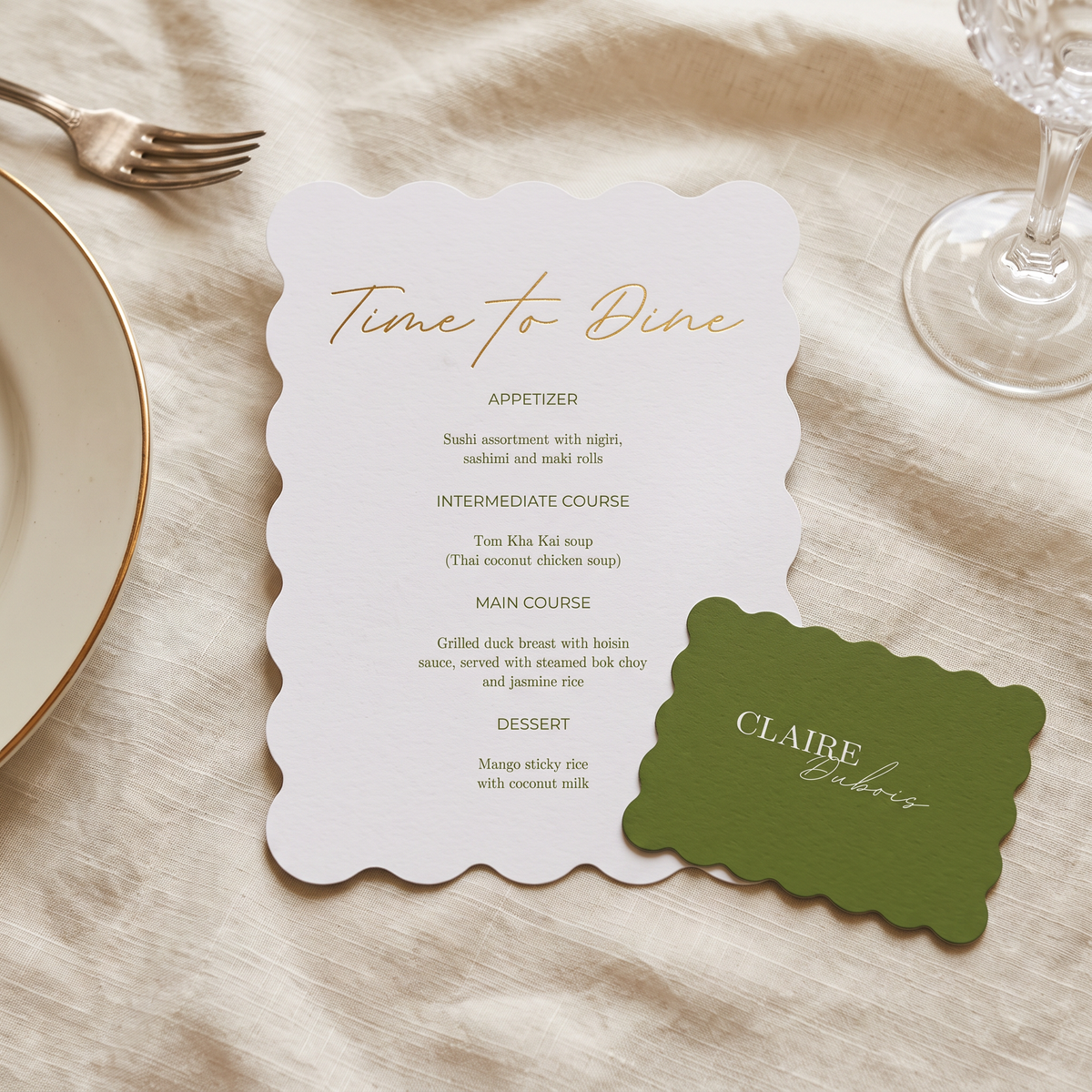 A scalloped white dinner menu card with gold script and printed course text, placed on a beige fabric surface with a green name tag reading 'CLAIRE Catering' and part of a gold-rimmed plate and glass visible around it.