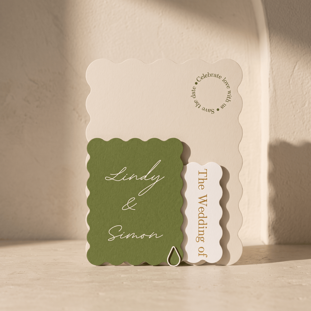 A wedding invitation suite featuring a scalloped cream card, an olive green card with white script reading 'Lindy & Simon,' and a small cream tab reading 'The Wedding of' on a warm beige surface.