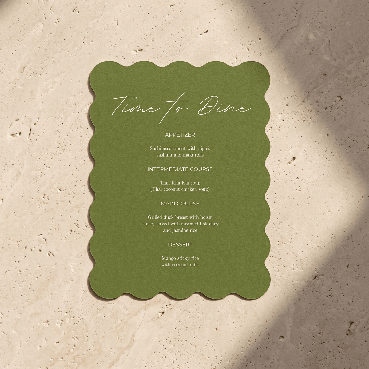 An olive green scalloped-edge menu card reading 'Time to Dine' with course text below, placed on a light beige stone surface with soft shadows.