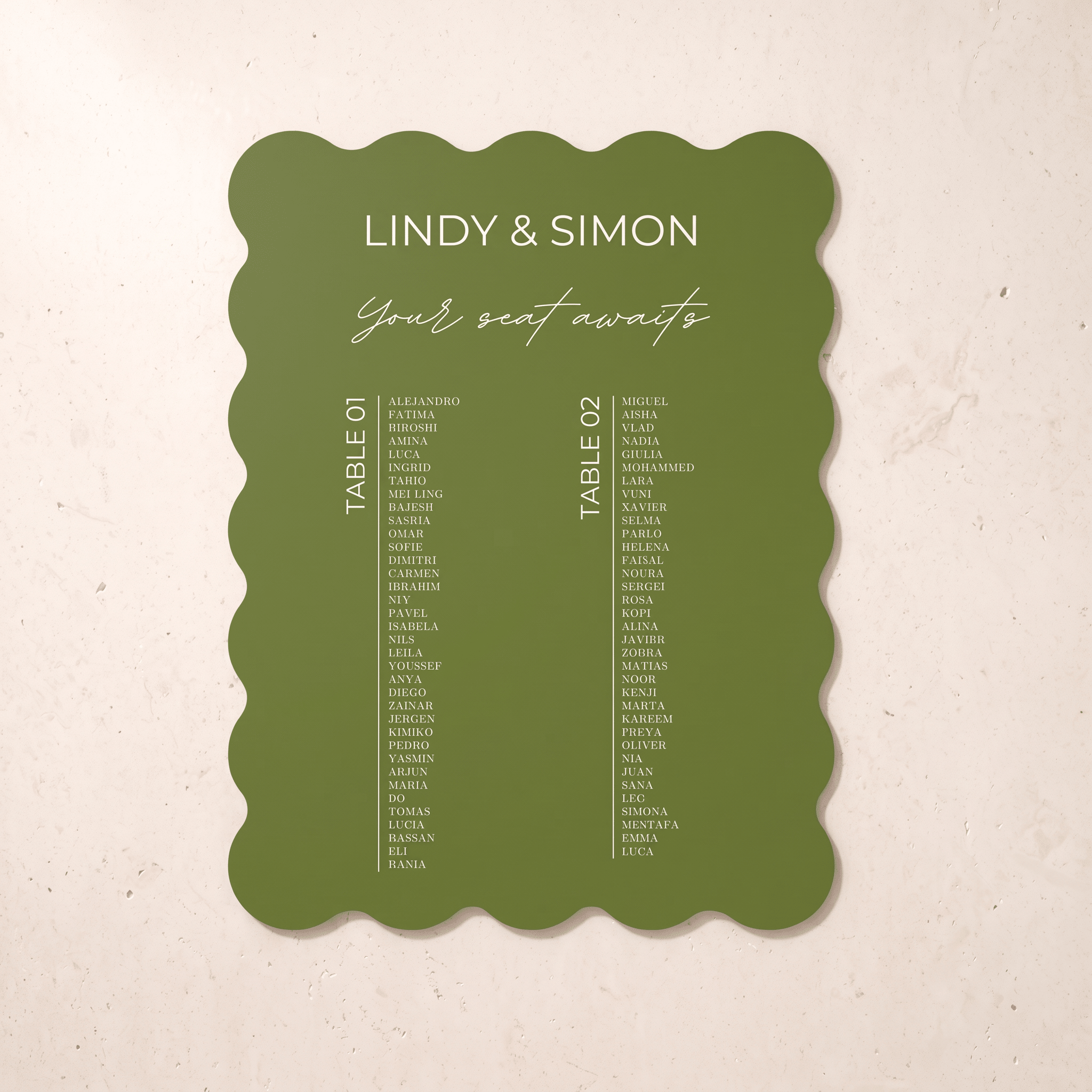An olive green scalloped-edge wedding seating card with white text reading 'LINDY & SIMON Your seat awaits' and two columns of guest names, photographed against a light beige textured background.