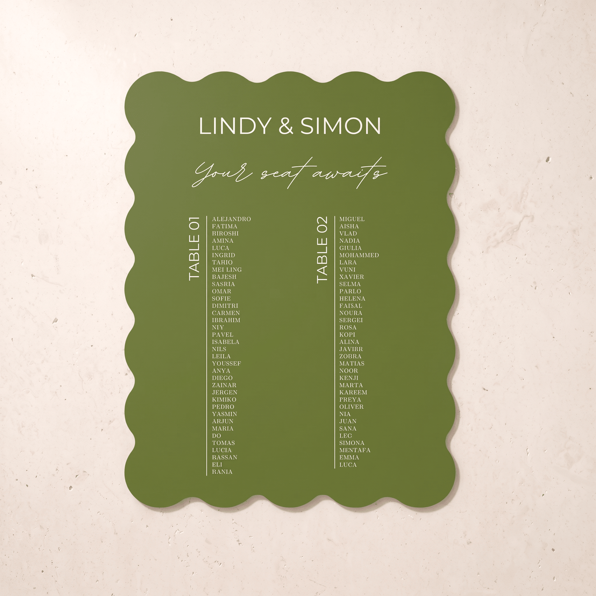 An olive green scalloped-edge wedding seating card with white text reading 'LINDY & SIMON Your seat awaits' and two columns of guest names, photographed against a light beige textured background.