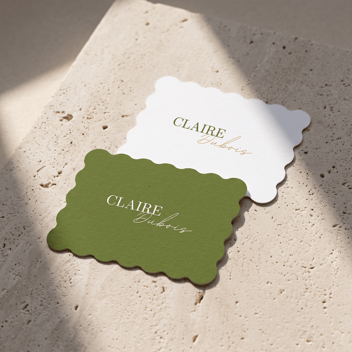 Two scalloped business cards for Claire Dubois, one in olive green and one in white, printed with gold foil text, arranged on a textured beige stone surface with soft diagonal shadows.