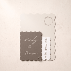 A wedding invitation suite featuring a taupe scalloped card with 'Lindy & Simon' in white script, layered over a cream card that reads 'Celebrate love with us' and a small white tag reading 'The Wedding of'.
