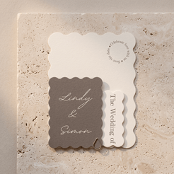 A coordinated set of wedding stationery cards with scalloped edges, including a taupe card reading 'Lindy & Simon,' an off-white card with script text, and a smaller off-white vertical card, arranged on a light stone surface.