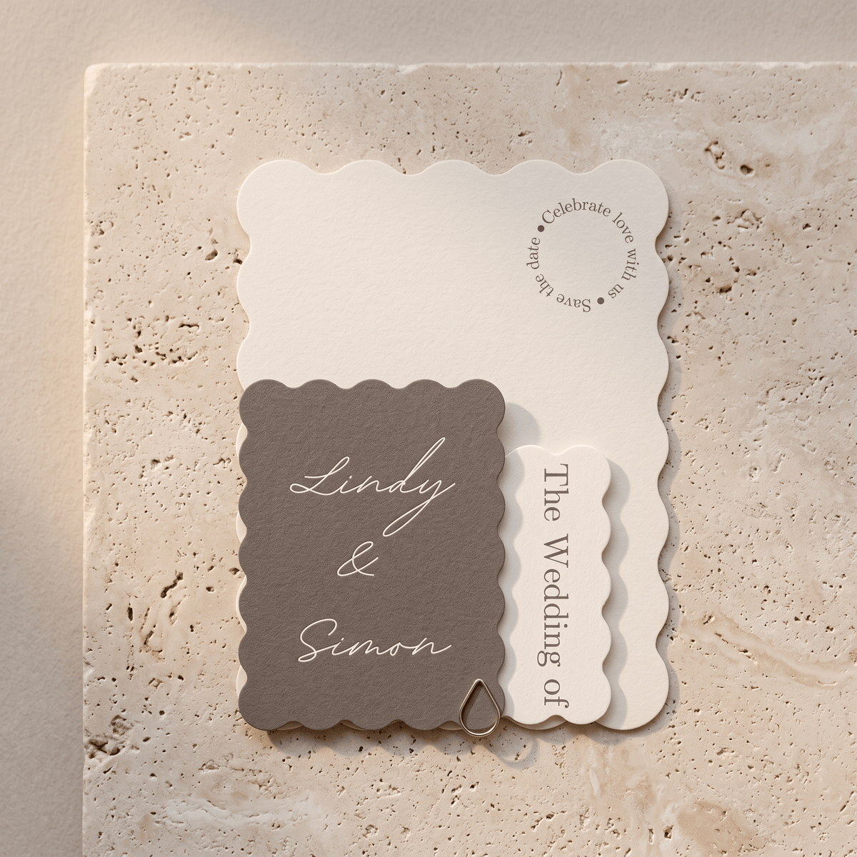A coordinated set of wedding stationery cards with scalloped edges, including a taupe card reading 'Lindy & Simon,' an off-white card with script text, and a smaller off-white vertical card, arranged on a light stone surface.