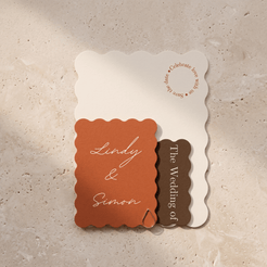A wedding invitation suite featuring a cream scalloped card, a terracotta orange scalloped card with white script reading 'Lindy & Simon', and a dark brown vertical tag reading 'The Wedding of', arranged on a beige stone surface.