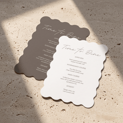 Two scalloped-edge paper menu cards with cursive 'Time to Dine' headings, one anthracite and one white, arranged on a textured beige stone surface with natural shadows.
