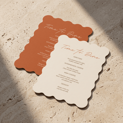 Two scalloped-edge paper menu cards on a textured stone surface, one terracotta orange and one warm cream, both printed with elegant script and course lists.