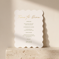 A scalloped-edge white menu card with elegant gold foil text reading 'Time to Dine' and course listings below, displayed on a beige stone pedestal against a warm cream wall.