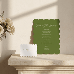 A green scalloped-edge menu card reading 'Time to Dine' stands on a stone ledge beside a small white card with the brand name 'CLAIBE dining' against a warm beige wall with soft window shadows.