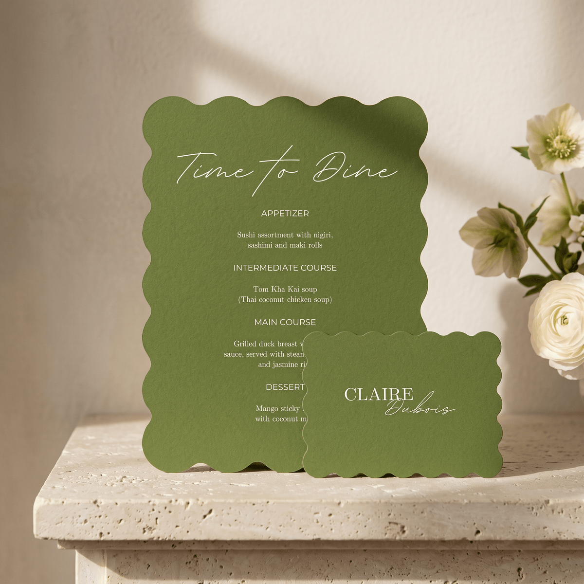 A green scalloped wedding menu card and matching place card on a stone surface, with white script text reading 'Time to Dine' and the names 'CLAIRE Dubois'.