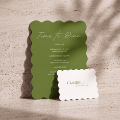 A green scalloped-edge restaurant menu card standing on a stone surface, with a white scalloped-edge name card in front reading 'CLAIRE bistro.'.
