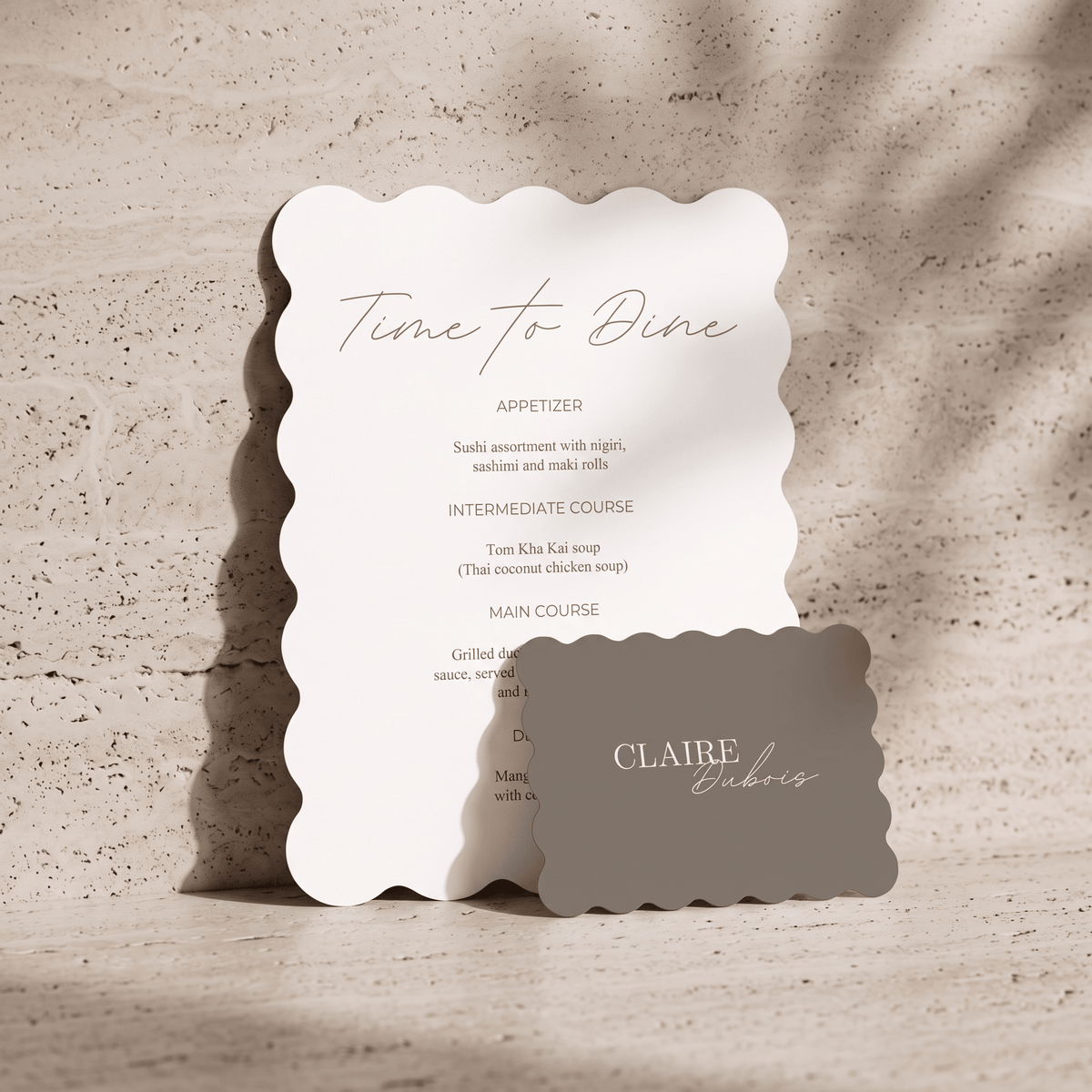 A scalloped-edge white dinner card with elegant script reading 'Time To Dine' and a anthracite place card reading 'CLAIRE & bistro' resting in front, set on a textured beige stone surface with soft leaf shadows.