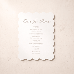 A scalloped white paper dinner menu card with elegant script and course text, photographed flat on a light beige textured surface.