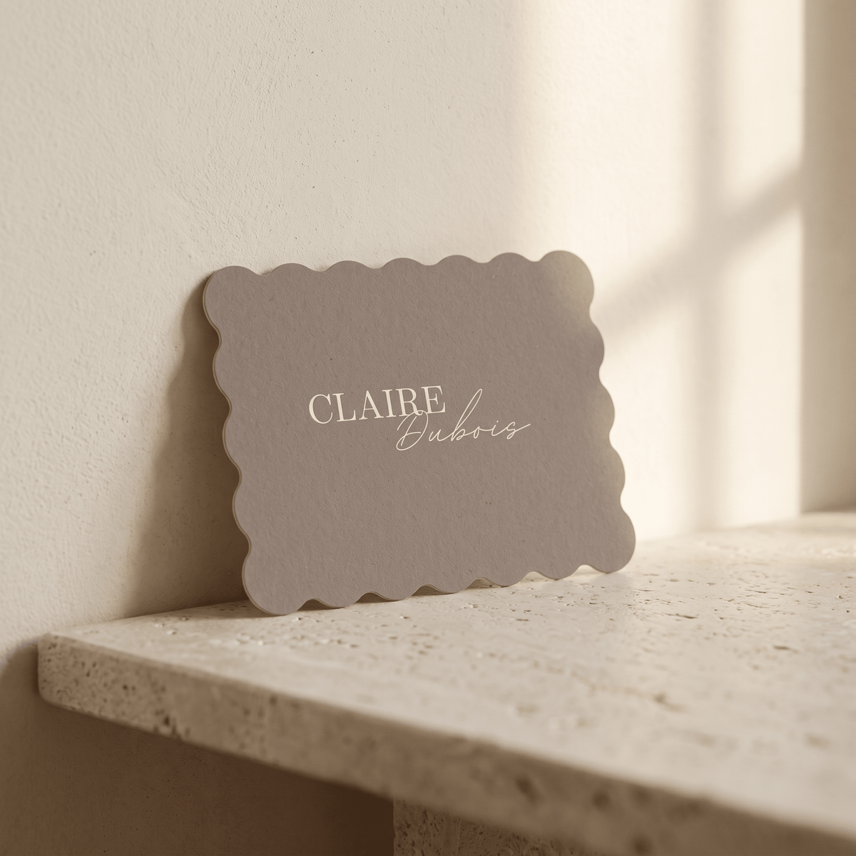 A scalloped-edge taupe paper card with the names 'CLAIRE Dubois' printed in white, leaning against a textured beige wall on a stone ledge.