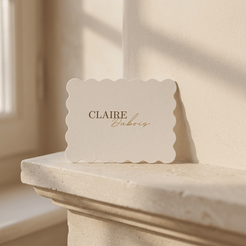 A cream-colored paper card with scalloped edges, printed with the name 'CLAIRE' in dark brown serif and 'Eubrios' in copper-gold cursive, resting on a stone mantelpiece in warm sunlight.