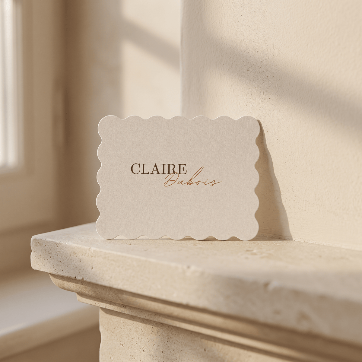 A cream-colored paper card with scalloped edges, printed with the name 'CLAIRE' in dark brown serif and 'Eubrios' in copper-gold cursive, resting on a stone mantelpiece in warm sunlight.