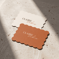 Two scalloped-edge paper cards on a light stone surface, one cream and one terracotta, both printed with the name Claire Cubais.