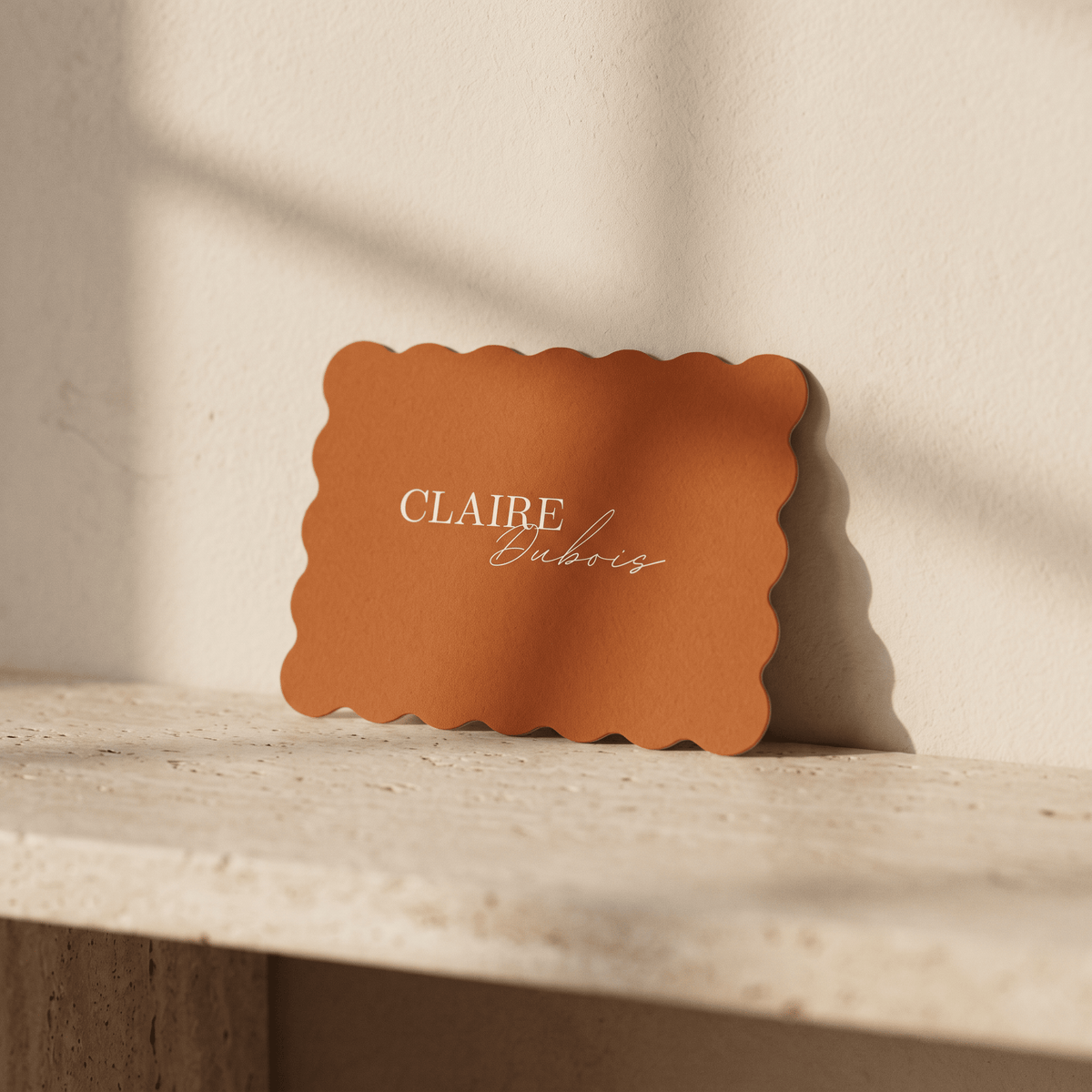 A rectangular terracotta-colored place card with scalloped edges and the text 'CLAIRE Dubois' printed in white, resting on a light stone ledge against a textured beige wall with soft window shadows.