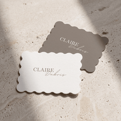 Two scalloped-edge paper cards printed with the name 'CLAIRE Dubois' in elegant serif and cursive typography, displayed on a textured beige stone surface with soft natural shadows.