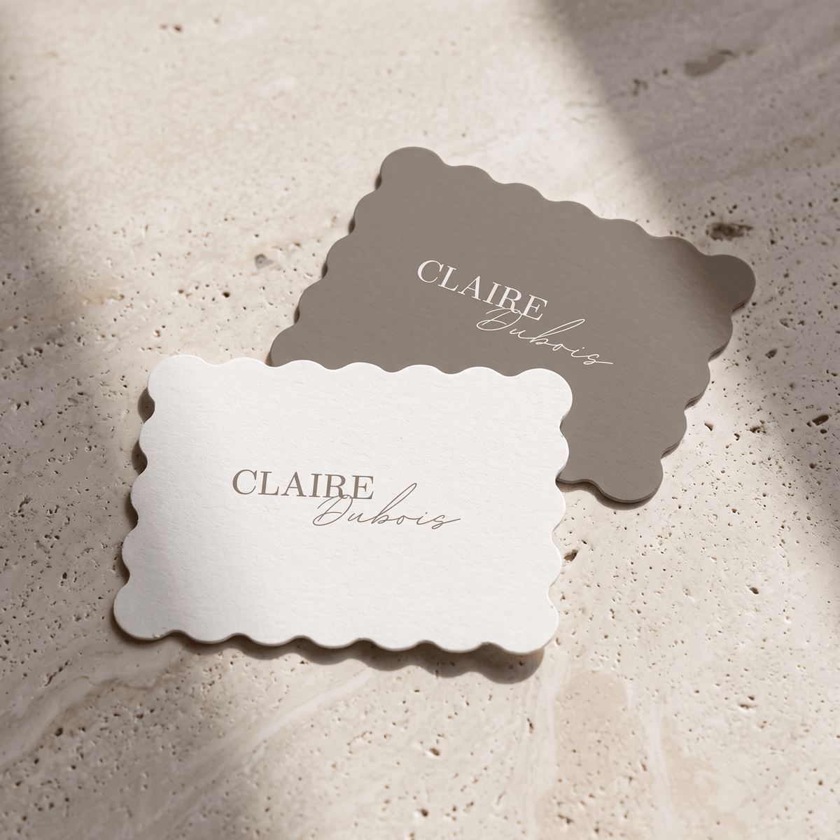 Two scalloped-edge paper cards printed with the name 'CLAIRE Dubois' in elegant serif and cursive typography, displayed on a textured beige stone surface with soft natural shadows.