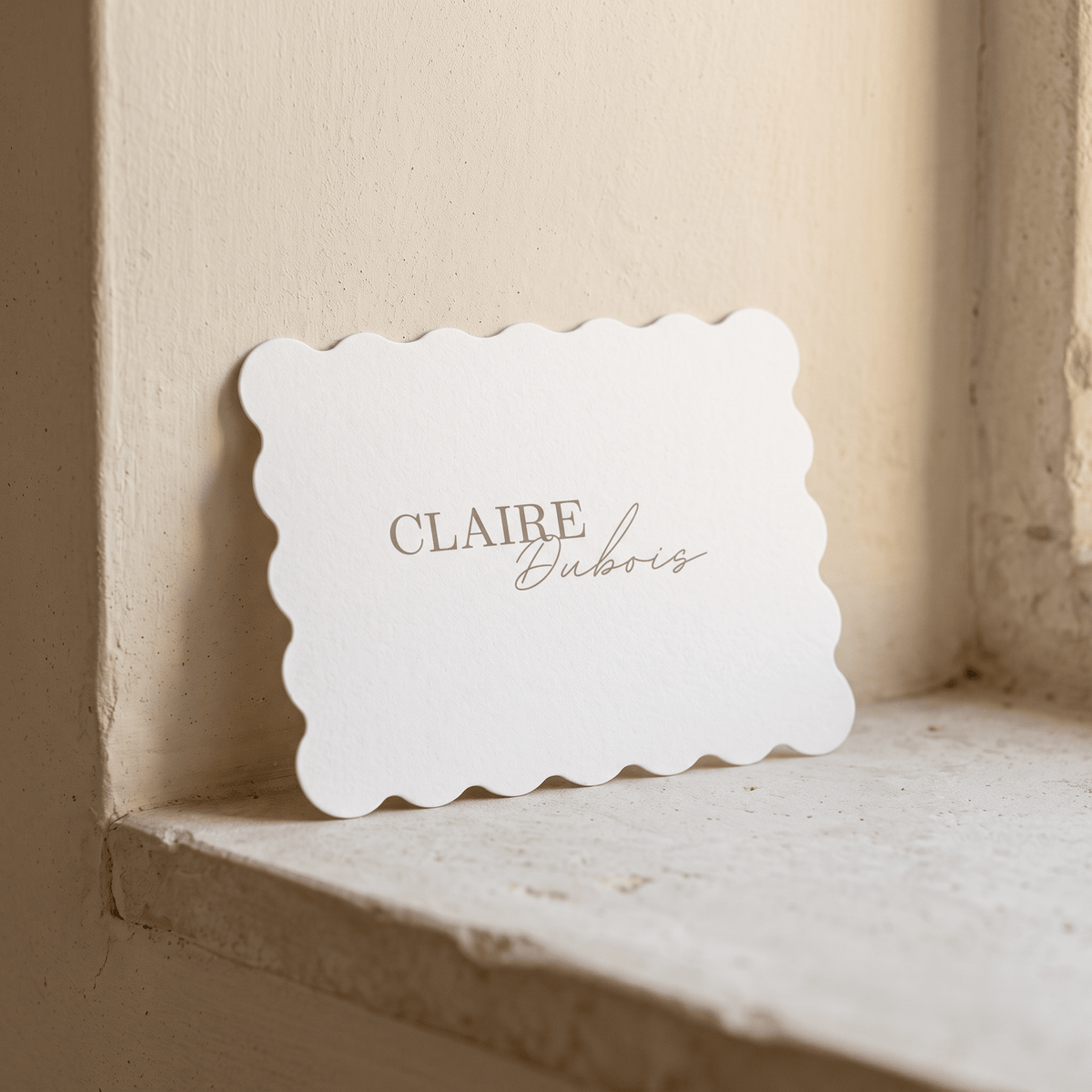 A white scalloped-edge card with the text 'CLAIRE Dubois' printed in a muted taupe, leaning against a textured beige wall on a stone ledge.
