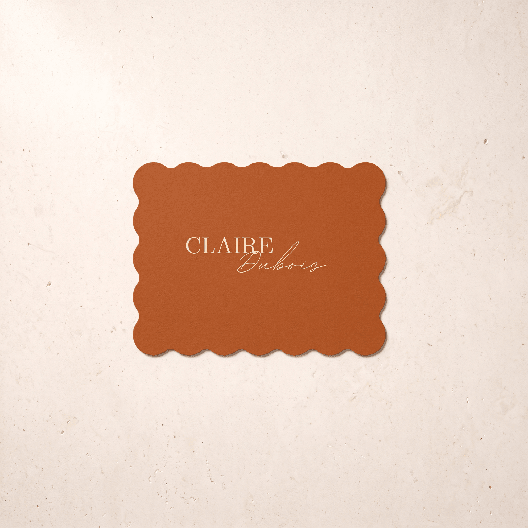 A rectangular terracotta-colored card with scalloped edges and the text 'CLAIRE Dubois' in gold, centered on a textured cream background.