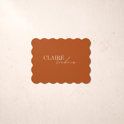 A rectangular terracotta-colored card with scalloped edges and the text 'CLAIRE Dubois' in gold, centered on a textured cream background.