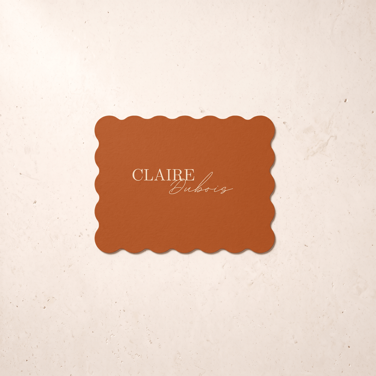 A rectangular terracotta-colored card with scalloped edges and the text 'CLAIRE Dubois' in gold, centered on a textured cream background.