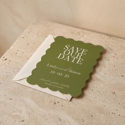 A scalloped olive green 'Save Our Date' card with white text, layered over a white envelope on a beige stone surface against a warm beige wall.
