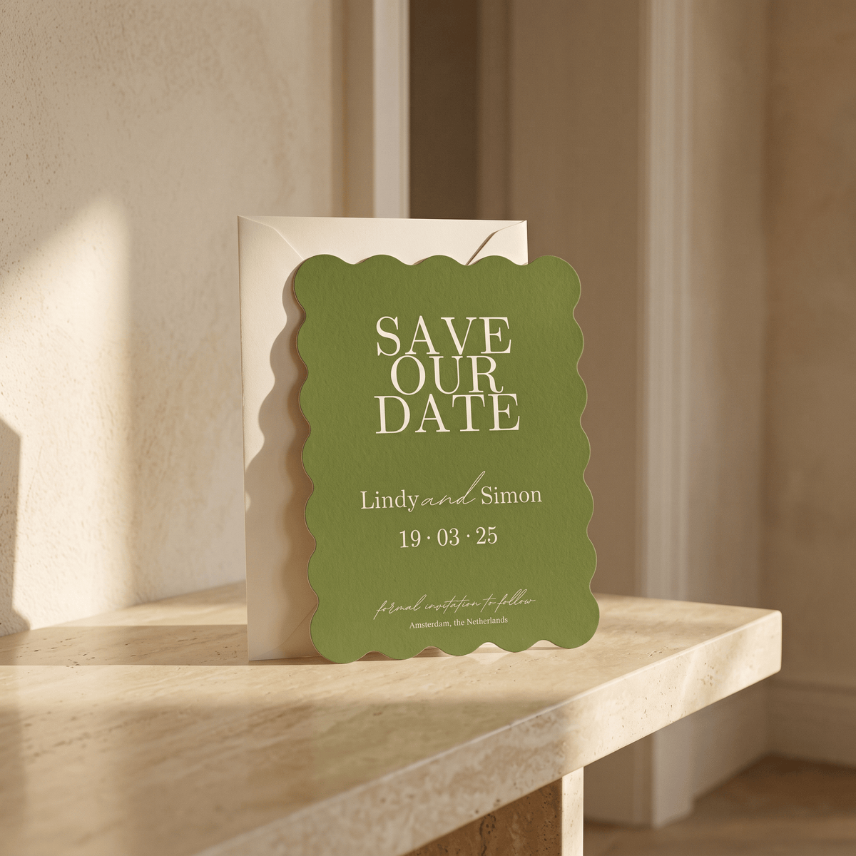 A green scalloped-edge 'Save Our Date' card with white text stands in front of a white envelope on a light wood table in a softly lit interior setting.