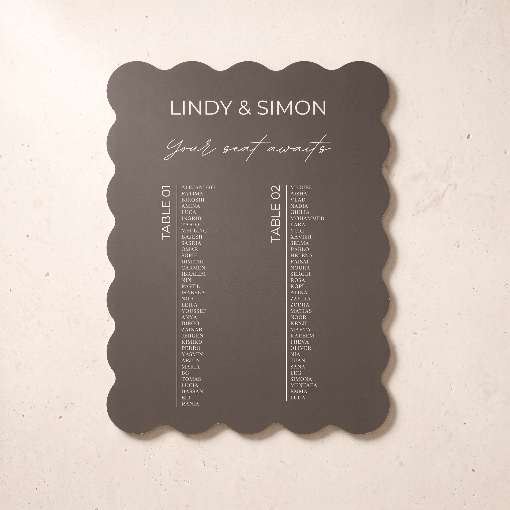 A scalloped anthracite wedding seating card with white printed text, displayed flat against a light beige textured background.