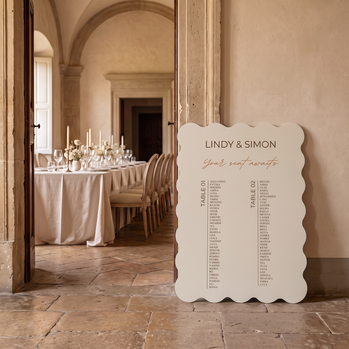 A scalloped-edge white wedding seating chart sign leaning against a wall in a rustic, elegantly decorated hall with stone floors and a table set in the background.