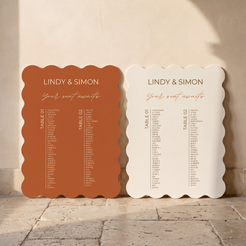 Two scalloped wedding seating cards displayed side by side on a stone floor against a beige wall. One card is terracotta orange and the other is warm off-white, both printed with the names 'LINDY & SIMON', 'Your seat awaits', and table list text.