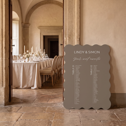 A scalloped-edge wedding seating sign in a warm taupe-gray color stands on a stone floor in a beautifully decorated event hall, with a table set visible through an open doorway in the background.