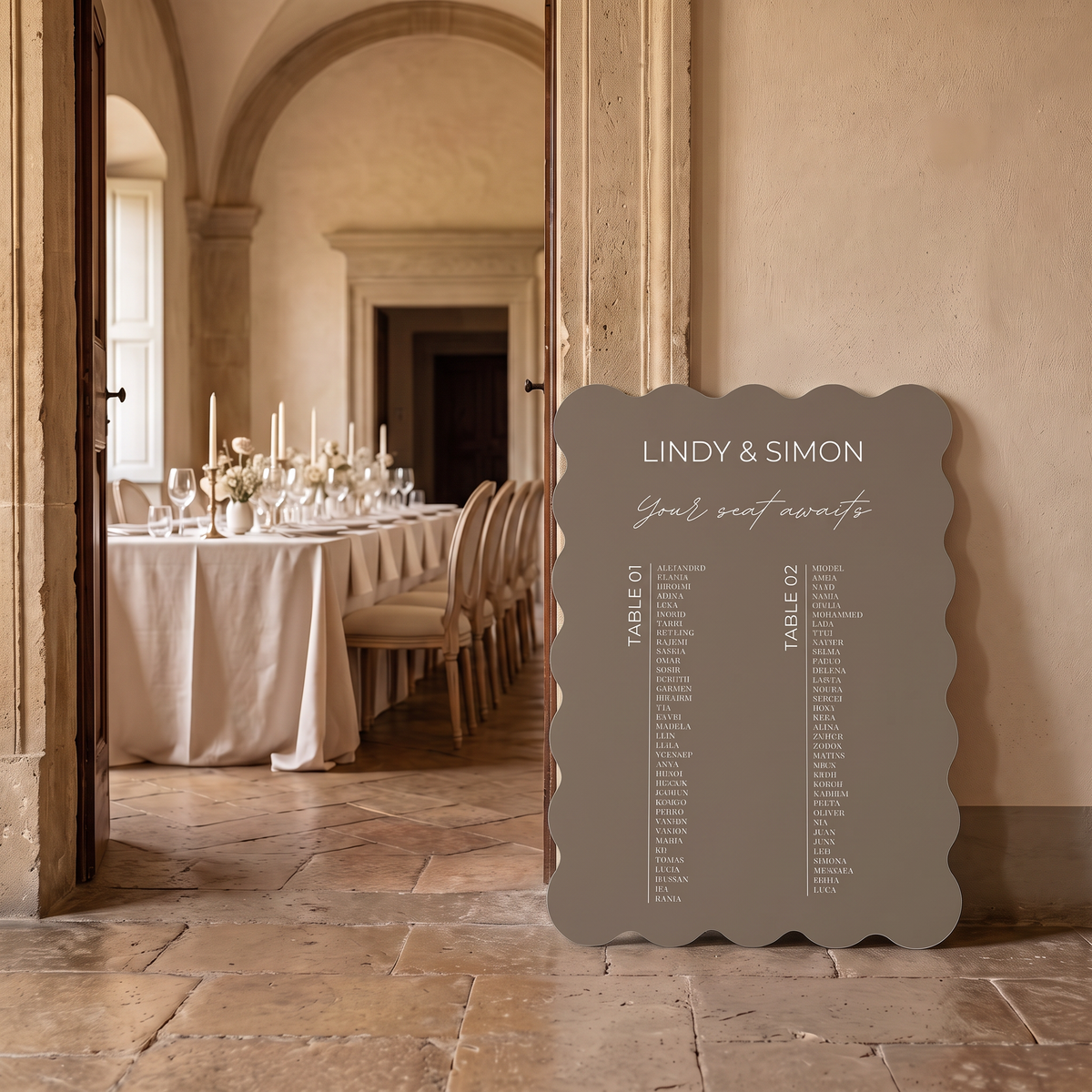 A scalloped-edge wedding seating sign in a warm taupe-gray color stands on a stone floor in a beautifully decorated event hall, with a table set visible through an open doorway in the background.