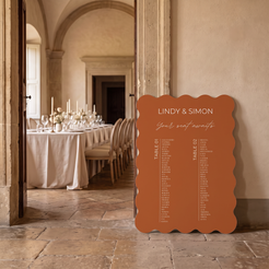 A scalloped terracotta wedding seating chart sign leaning against a stone wall in an elegant dining room setting.